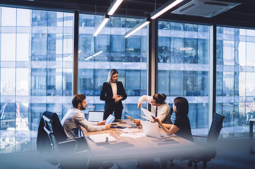 High angle of businesswoman in classy suit standing anear table in front of group of colleagues and checking time on wristwatch while working together on business project